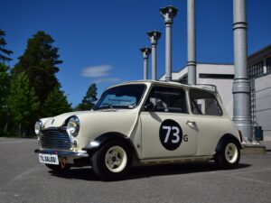 A classic cream-colored Mini Cooper with the number "73G" on its door, parked outside on a sunny day with trees and industrial columns in the background.