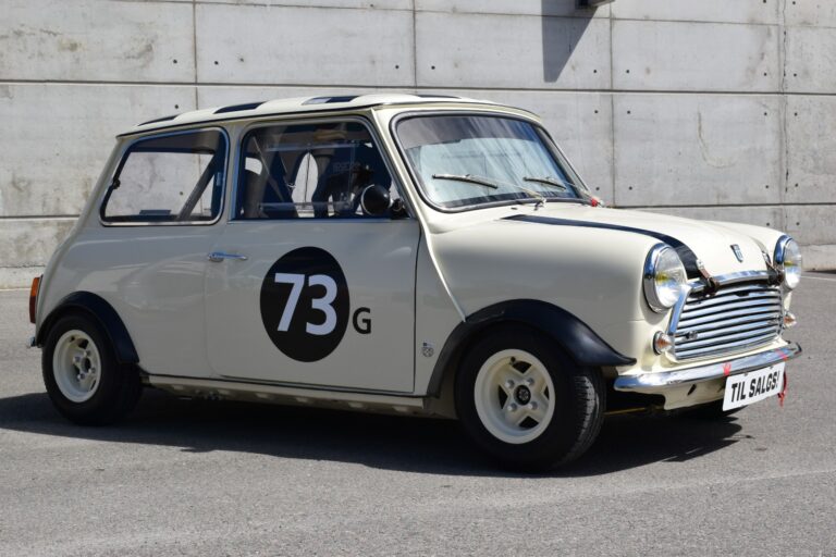 A vintage white Mini Cooper car with number 73G on its side, parked on a street against a concrete wall. The car has a racing setup with various modifications and unique license plate "TIL 5145B.