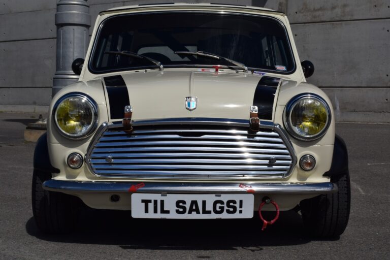 Front view of a vintage white car with black racing stripes and a "TIL SALGS!" sign on the front bumper, indicating it is for sale. The background includes a concrete wall and a light pole.