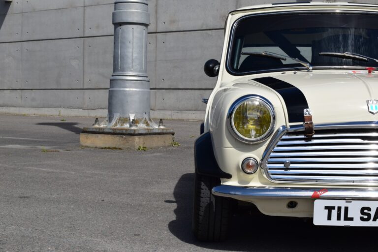 A cream-colored classic Mini Cooper with a black stripe is parked in front of a concrete wall and large metal pole. The license plate reads "TIL SALG.