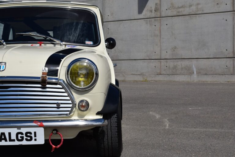 Front view of a classic white car with yellow headlights, black racing stripes, and a "HALGS!" license plate, parked in front of a concrete wall.