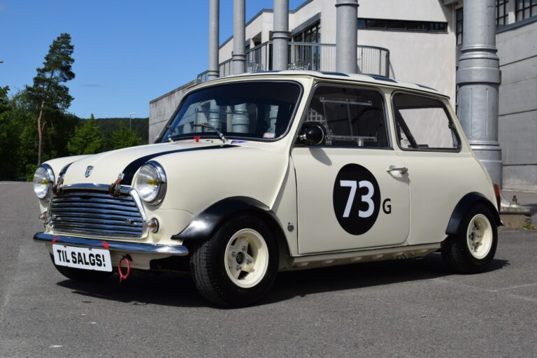 A vintage cream-colored Mini Cooper with racing number 73 displayed on the door, parked outdoors with a "Til Salgs!" sign on the front bumper.