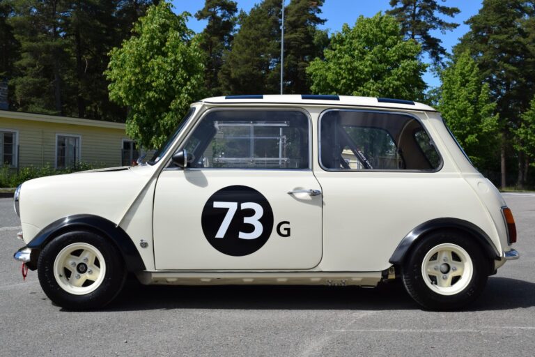 A side view of a cream-colored vintage mini car with the number 73 and the letter G on the door, parked outdoors near trees and a building.