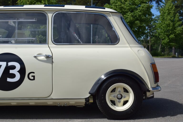 Side view of a cream-colored vintage Mini Cooper with the number "73G" painted on its door, showing the rear half of the car, including the wheel and part of the window.