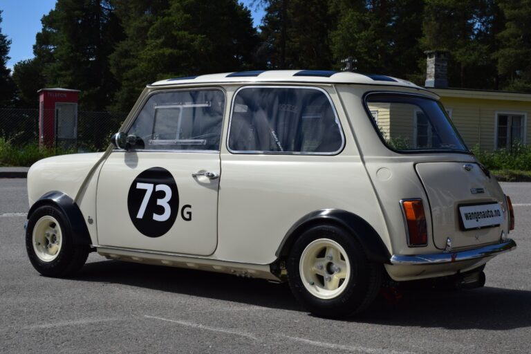 A white vintage Mini Cooper with the number 73G on the door, parked outdoors on a sunny day with trees and a building in the background.