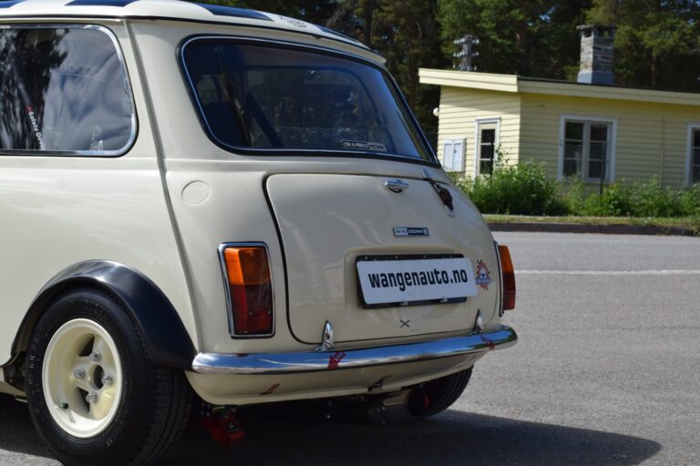 Rear view of a cream-colored vintage car with a "wangenauto.no" sticker on the license plate, parked outdoors near a small yellow building surrounded by greenery.