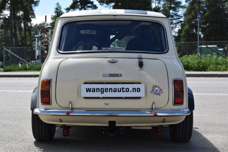 Rear view of a cream-colored vintage Mini Cooper S parked on a street, with a visible "wangenauto.no" license plate and various stickers on the rear window.