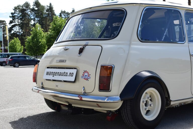 Rear view of a vintage beige Mini Cooper parked outdoors with a "wangenauto.no" website decal on the rear bumper. Trees and cars are visible in the background.