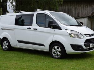 White Ford Transit van parked on grass in front of a wooden building with trees in the background.