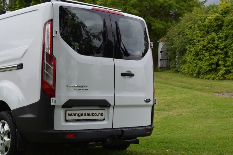 White van parked on grass with rear doors showing, featuring "wangenauto.no" branding. Trees and a building visible in the background.