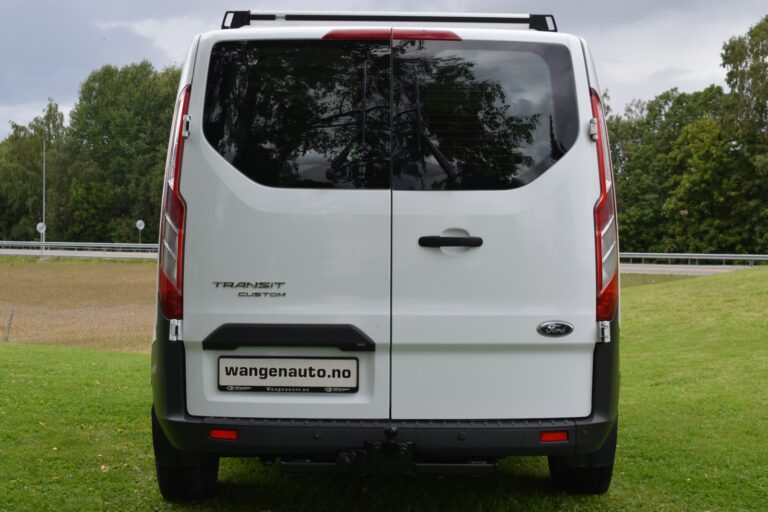 A white Ford Transit Custom van is parked on grass, shown from the rear with a "wangenauto.no" license plate. Trees and a road are visible in the background.