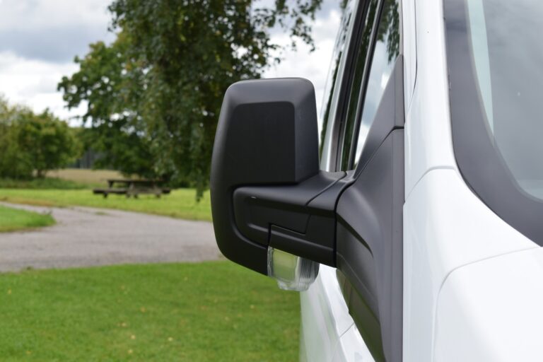 Close-up of a black side mirror on a white vehicle with green grass, trees, and a picnic table in the background.