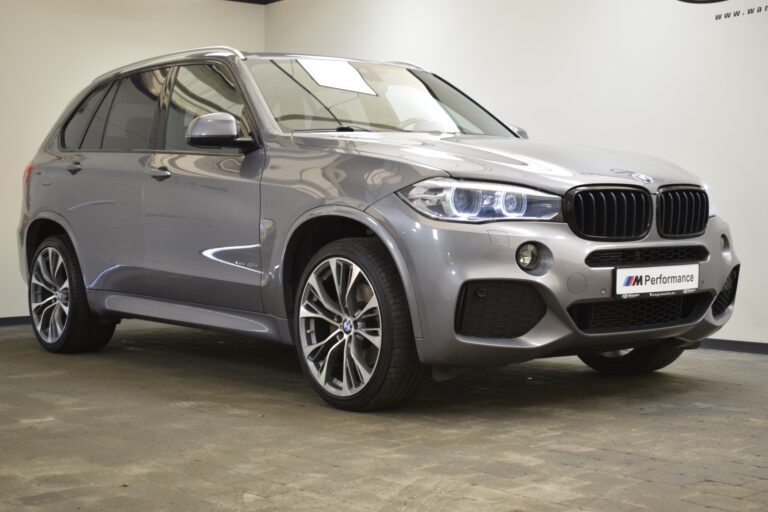 A silver BMW SUV is parked in a showroom with a "M Performance" plate on the front. The vehicle is viewed from a three-quarter front angle.