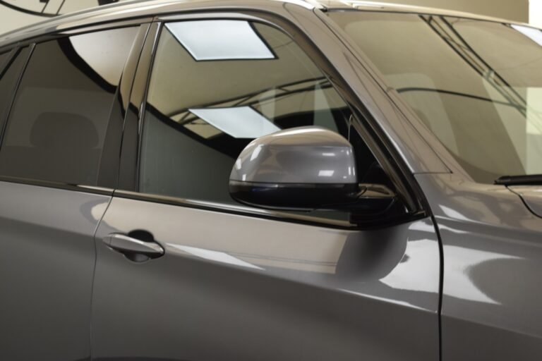 Close-up of the side of a gray car showing the driver's side door, window, and side mirror, against a background of ceiling lights.