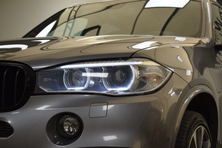 Close-up of the front left side of a grey car, showing the headlight, part of the grille, and a fog light below. The headlights are on, and the car has a sleek, modern design.