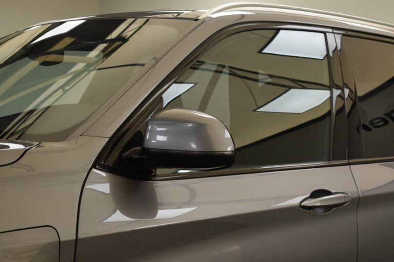 Close-up of a car’s side mirror and part of the front door featuring tinted windows, captured in an indoor setting with reflections of overhead lights visible on the vehicle.