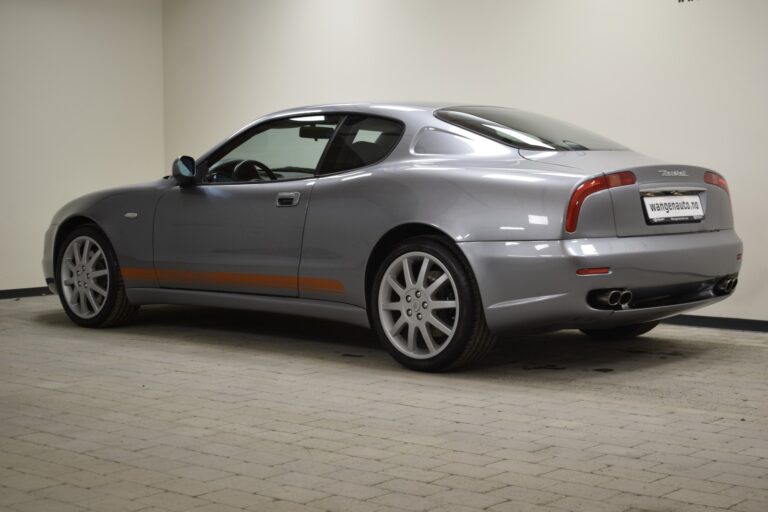 A silver two-door sports car is parked indoors on a tiled floor, with dual exhaust pipes visible at the rear. It has a sleek design and alloy wheels.