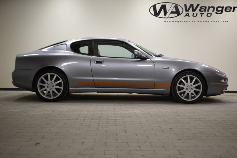 A silver coupe with orange accents on a tiled floor, parked indoors. The wall in the background displays the "Wanger Auto" logo and website URL.