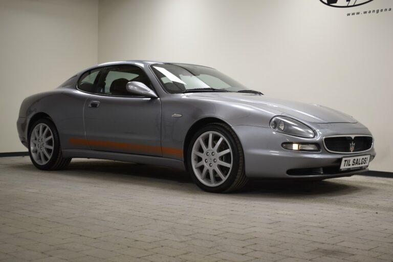 A silver Maserati coupe is parked indoors on a tiled floor, with a custom license plate that reads "TIL SALGS." The car has a sleek design and is photographed from the front-left angle.