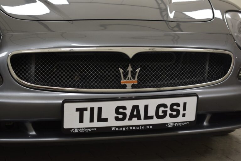 Close-up of a car's front grille with a license plate that reads "TIL SALGS!" and a website "Wangenauto.no" displayed beneath it. Vehicle is metallic gray with a distinctive trident emblem.