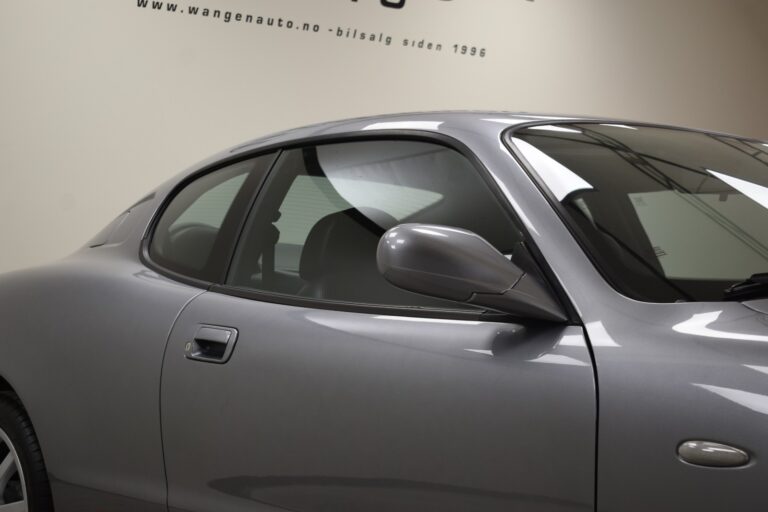 A close-up view of a gray car’s side, highlighting the door, side mirror, and partially tinted windows in a showroom. The website "wangenauto.no" is visible in the background.