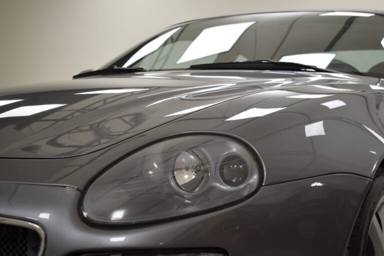 Close-up of a sleek, shiny grey sports car, focusing on the headlight and hood area. The car's exterior reflects the lighting above.