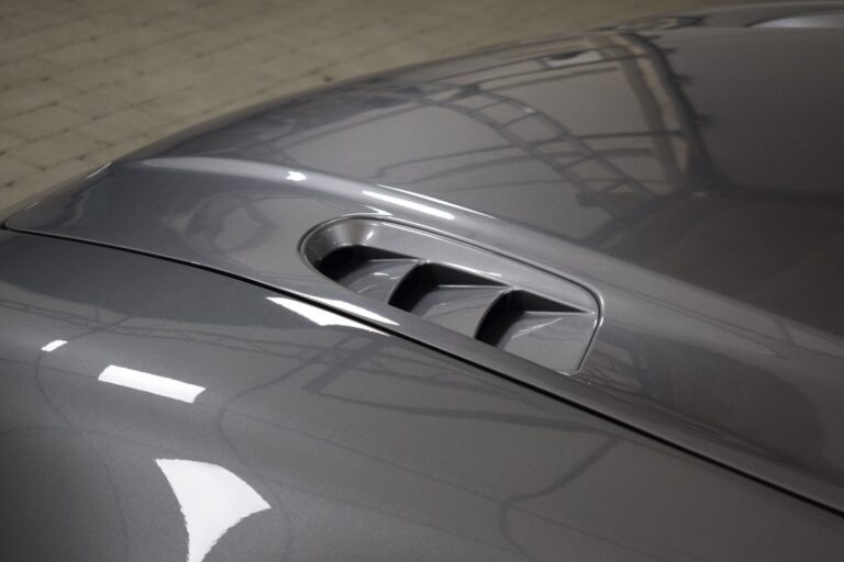 Close-up of a silver car's hood, showing an air vent with three slats. The surface has a glossy finish, reflecting overhead lighting and nearby structures. The background reveals a tiled floor.