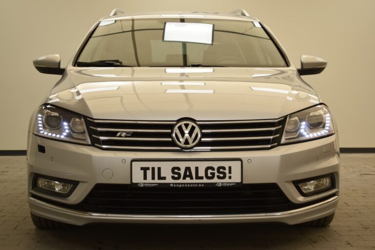 Front view of a silver Volkswagen car with illuminated headlights and a sign on the license plate area that reads "Til Salgs!" (For Sale!). The background is a plain indoor setting.