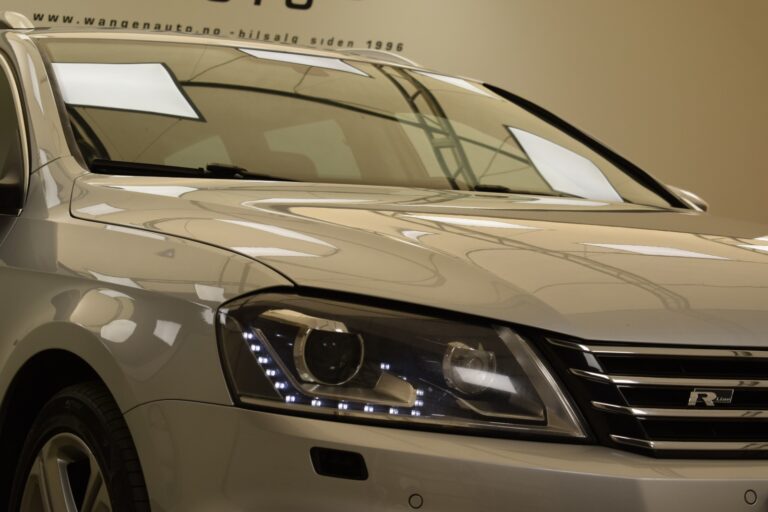 Close-up of the front left side of a silver car showing the headlight, grille, and part of the hood and windshield. The background includes part of a wall with some text visible at the top.