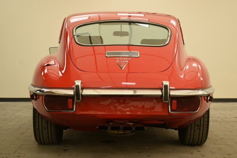 Rear view of a red vintage Jaguar E-Type car, showcasing the chrome bumper, dual exhaust pipes, and distinctive tail light design against a plain background.