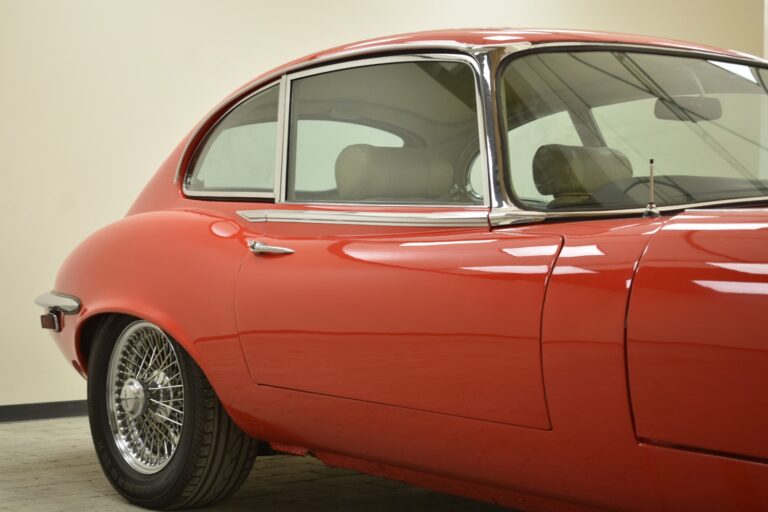 Side view of a red classic car with a curved roof and wire-spoked wheels, parked indoors.