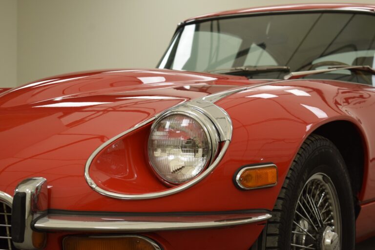 Close-up of the front end of a vintage red sports car, highlighting the headlight, chrome details, and part of the windshield.