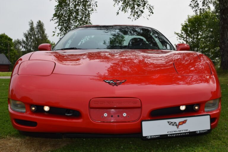 A red Corvette sports car is parked on a grassy area, with the front view prominently displaying the Corvette emblem on the hood. Trees and part of a house are visible in the background.