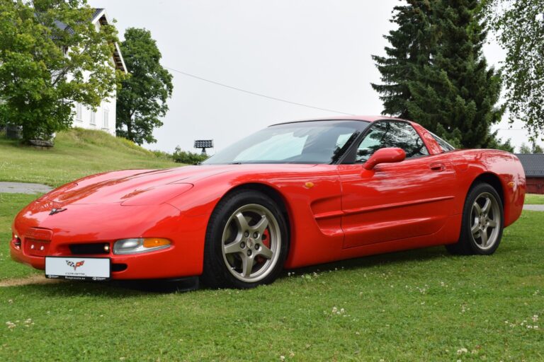 A red sports car is parked on grass with trees and a house visible in the background.