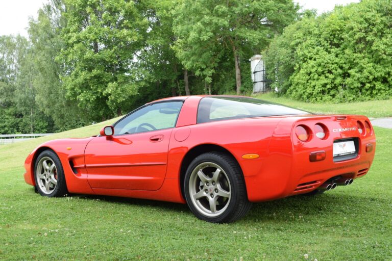 A red sports car is parked on a grassy area with trees in the background. The car has a sleek design, dual exhausts, and is labeled as a “Corvette” on the back.