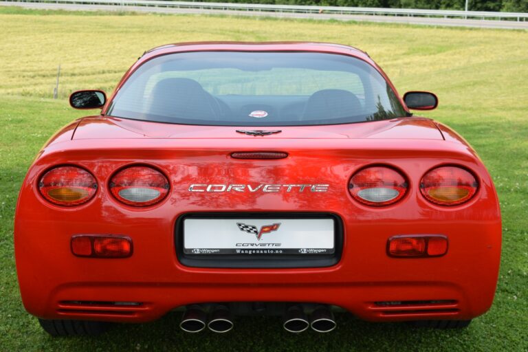 A red Corvette sports car is parked on a grassy field, viewed from the rear. The car features dual exhaust pipes, round taillights, and the Corvette logo and branding on the back.