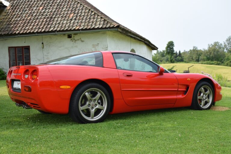 A red sports car is parked on a grassy area near a rustic house with a tiled roof.