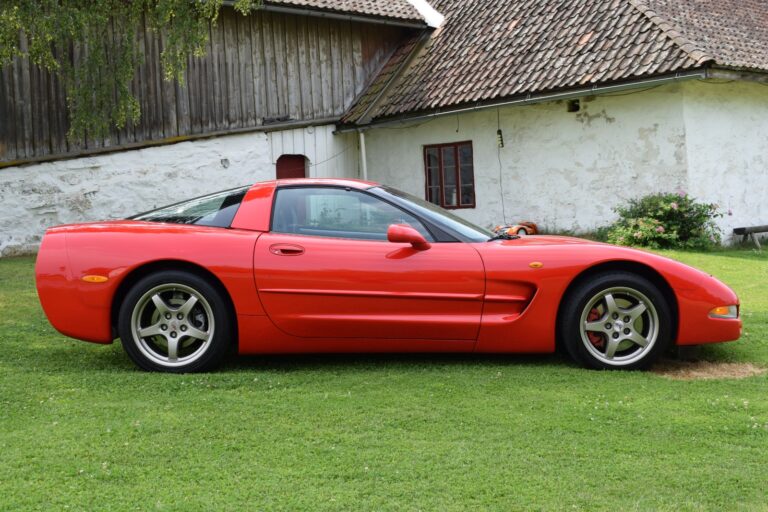 A red sports car is parked on a grassy area in front of a white, rustic building with a wooden roof and framed windows.