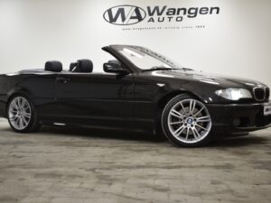 A black convertible car with its roof down, parked indoors under signage that reads "Wangen Auto." The car is facing left with reflective lighting on its polished surfaces.