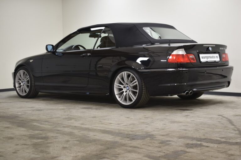 A black convertible car with a soft top roof parked in an indoor setting. The car is viewed from the rear side and has alloy wheels.