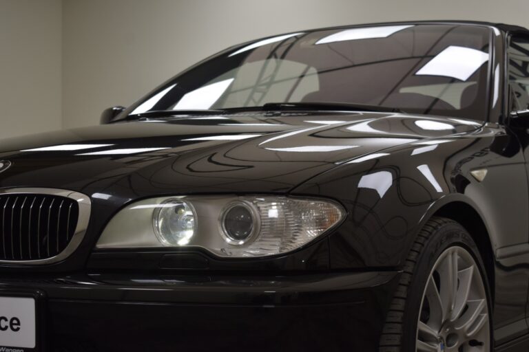 A close-up view of a black luxury car, focusing on its front left headlight and part of the grille, reflecting the indoor lighting on its polished surface.