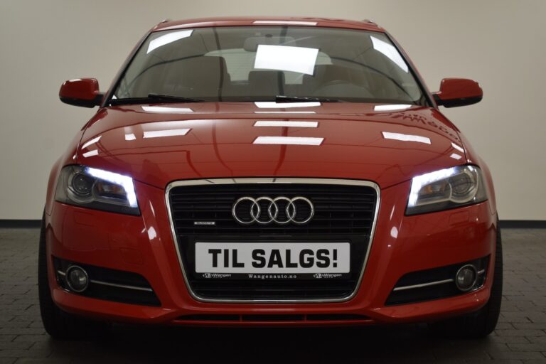 Front view of a red Audi car with headlights on, displayed indoors with a sign on the front bumper reading "TIL SALGS!" which means "FOR SALE!" in Norwegian.