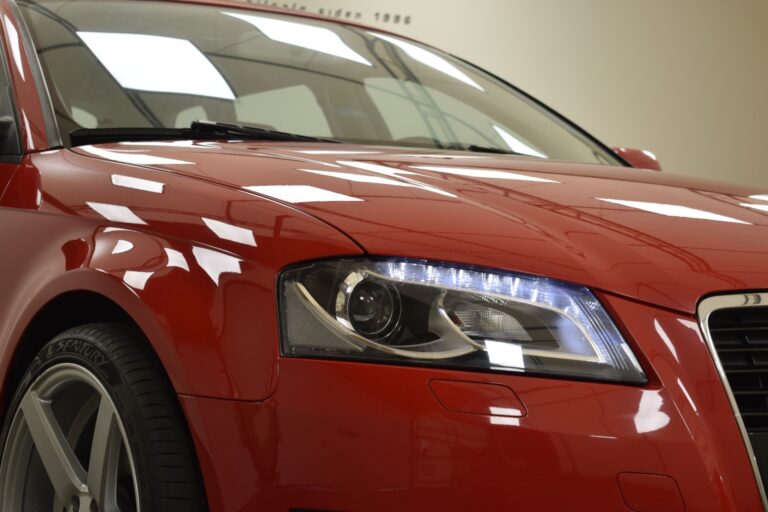 Close-up of the front end of a red car, highlighting the headlight, grille, and part of the front wheel. Indoor lighting is reflected on the car's surface.