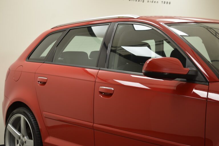 A close-up view of a shiny red car's side, showing the door, windows, side mirror, and rear wheel.