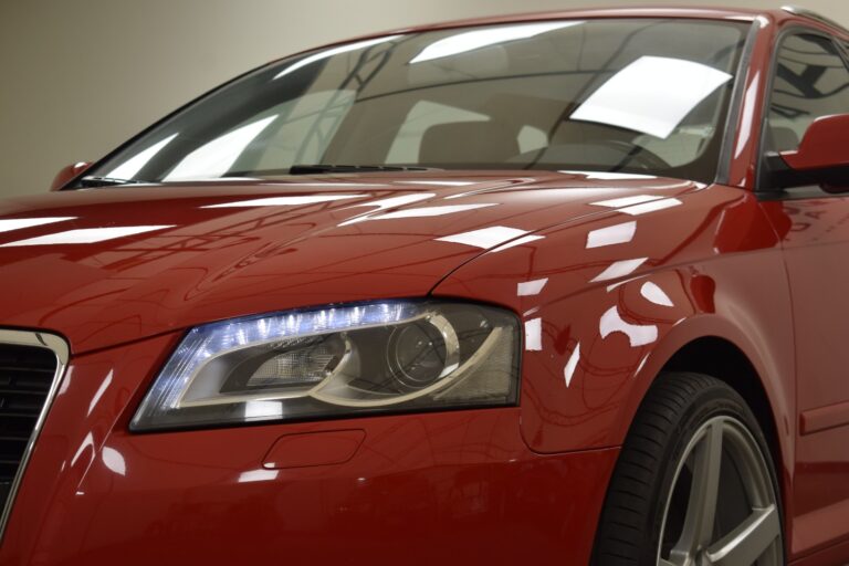 Close-up view of the front of a red car, focusing on the left headlight and part of the windshield and hood, reflecting indoor lighting.