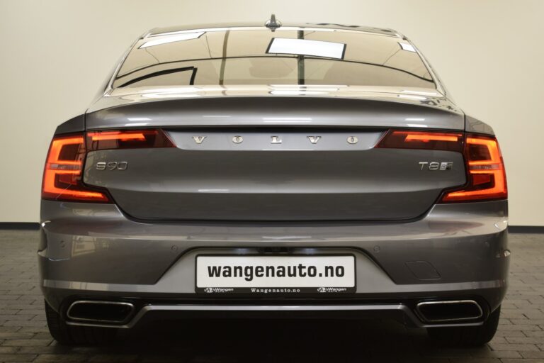 Rear view of a gray Volvo S90 T8 with illuminated tail lights, displayed in an indoor showroom. The car's license plate reads "wangenauto.no".