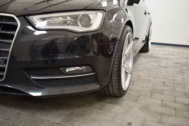 Close-up of a black car's front left corner, focusing on the headlight, bumper, and a portion of the tire, parked on a tiled floor.