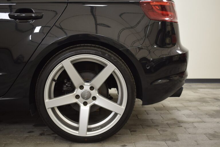 Close-up of the rear wheel and bumper of a black car, featuring a five-spoke alloy rim and a low-profile tire. The background includes a tiled garage floor and a plain wall.