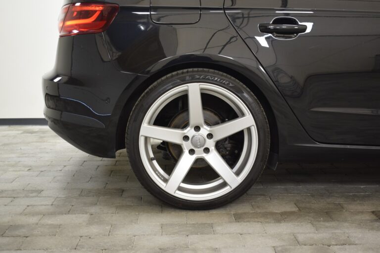 Close-up of the rear half of a black car showing the back wheel with a silver alloy rim and a visible brake disc, parked on a tiled surface.