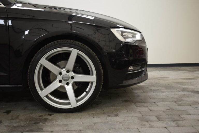 Close-up of the front left wheel and headlight of a shiny black car parked on a tiled floor.